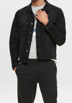 Mand iført sort denimjakke over lysegrå grafisk t-shirt og sorte bukser, justerer jakke kraven med en hånd.
