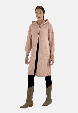 Mujer con un largo cárdigan rosa de punto con capucha, leggins negros y botas beige hasta la rodilla, de pie frente a un fondo liso.