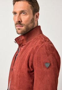Chaqueta de ante rojo con un alto cuello, que presenta un parche con el logo en el hombro izquierdo. La textura suave y el corte entallado realzan su diseño.