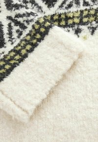 Détail d'un pull en tricot avec une texture crémeuse douce et duveteuse, présentant un motif géométrique noir, blanc et vert le long des poignets.