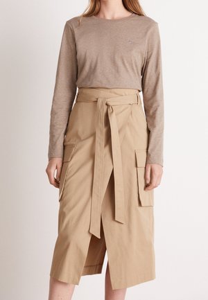 Vrouw draagt een beige top met lange mouwen en een hoge taille kaki rok met een voorste split, riem en grote zijzakken.
