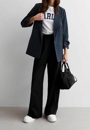 Nainen pukeutunut tummansiniseen bleiseriin, valkoiseen paitaan, jossa teksti "GIRL", mustiin leveälahkeisiin housuihin, valkoisiin lenkkareihin ja kantaa mustaa kangaskassia.