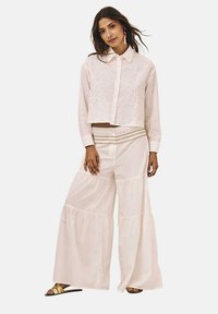 Camisa blanca corta con bordado en la parte delantera y el cuello, combinada con pantalones anchos blancos que presentan volantes en capas y una cinturilla decorativa.