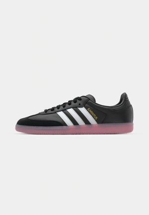 Schwarze Sportschuhe mit weißem Dreistreifen-Design, Wildlederakzenten, abgerundetem Zehenbereich und einer durchsichtigen pinken Gummisohle. Goldenes "JUVENTUS"-Logo.
