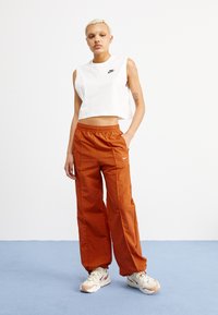 Top blanco recortado sin mangas y pantalones sueltos naranjas con cinturilla elástica. Logo de Nike en la parte superior y en el lado de los pantalones. Zapatillas de color claro.