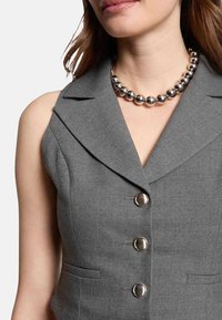 Gilet grigio senza maniche con colletto a P, dotato di quattro bottoni metallici rotondi e tasche laterali; texture liscia. Collana di perle argentate.