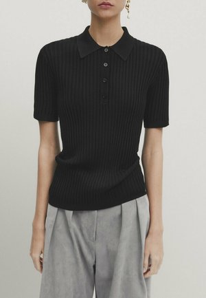 Donna che indossa una polo nera a maniche corte con coste abbinata a pantaloni grigi larghi su uno sfondo chiaro e semplice.