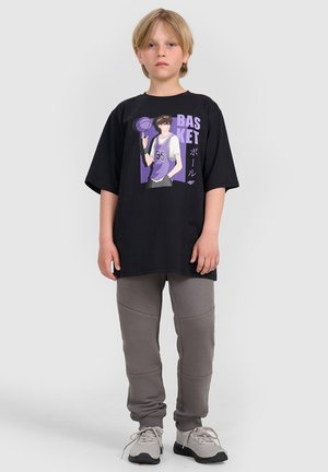Blond dziecko stoi, nosząc czarny oversize t-shirt z fioletową grafiką koszykówki, szare spodnie dresowe i jasnoszare trampki na białym tle.
