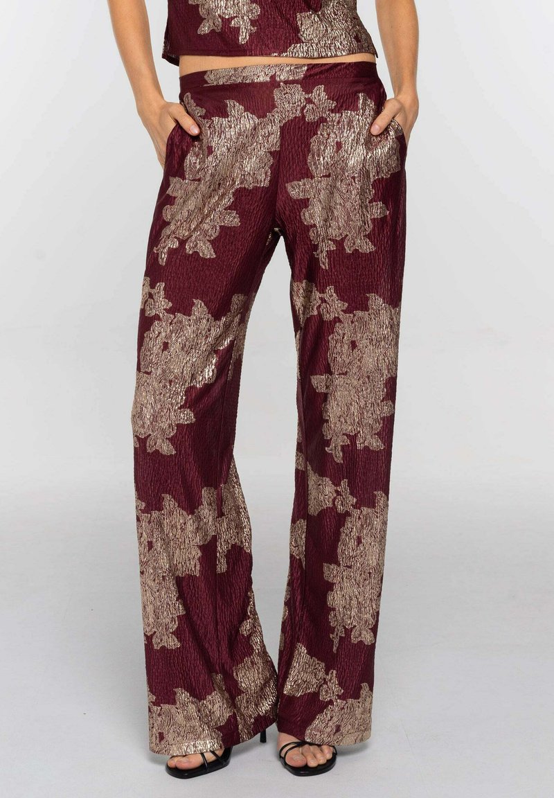 Mujer vestida con pantalones de pierna ancha burdeos con grandes patrones florales dorados y un crop top a juego, con las manos en los bolsillos y tacones abiertos negros visibles.