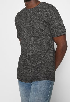 Ein Mann trägt ein dunkelgraues, strukturiertes Kurzarm-T-Shirt und hellblaue Jeans, während er vor einem schlichten weißen Hintergrund steht.