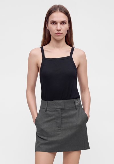 Femme aux longs cheveux raides, portant un haut noir sans manches et une jupe grise, les mains dans les poches, face à un fond uni.