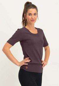 Lyhythihainen violetti paita pehmeästä kankaasta, pyöreällä kaula-aukolla ja kerroksilla helman kohdalla, jossa on oranssi logo. Mustat housut.