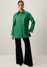 Zaļš mākslīgās ādas pārmērīgi liels krekls ar punktotu apkakli, lieliem pogām un platām aprocēm, apvienots ar melniem platām biksēm.