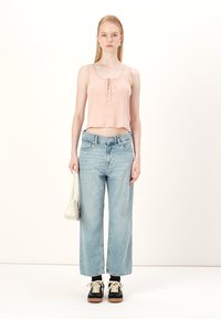 Top court rose pâle avec des boutons, associé à un jean bleu clair ample. Le mannequin tient un petit sac à main blanc, porté avec des baskets noires.