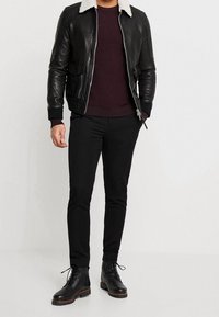 Czarna skórzana kurtka bomber z białym kołnierzem z owczej wełny, zestawiona z bordowym swetrem, czarnymi slim fit spodniami i czarnymi sznurowanymi butami.