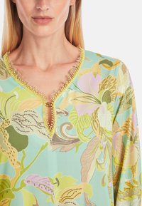 Blumen V-Ausschnitt Bluse in Hellgrün mit goldenen Akzenten, die eine Vielzahl von pastellfarbenen botanischen Mustern und einem weichen, leichten Stoff aufweist.