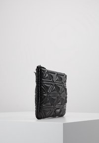 Černá prošívaná taška s geometrickým trojúhelníkovým vzorem na bílé podstavě na jednobarevném světlém pozadí.