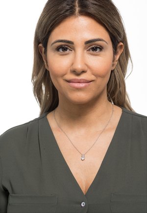 Collana in argento con un piccolo pendente rotondo con due pietre trasparenti, indossata su una blusa verde con scollatura a V e dettaglio tasca.