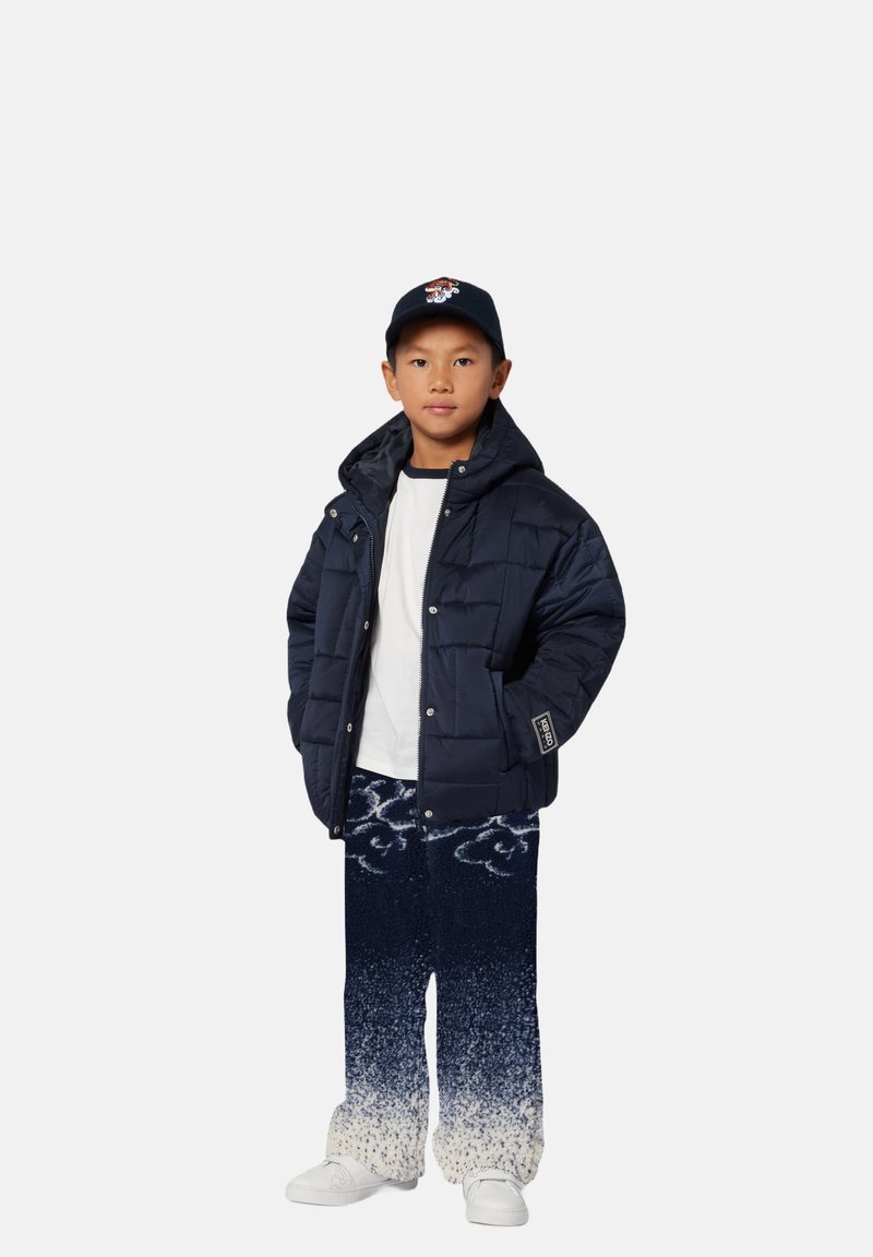 Child wearing a navy puffer jacket, white shirt, and patterned blue pants, standing confidently with a baseball cap and white sneakers.