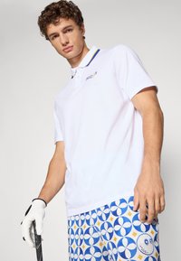 Hvit polo skjorte med en krave i blå og gul striper, med en liten brodert logo. Kombinert med mønstrede blå og gule shorts.