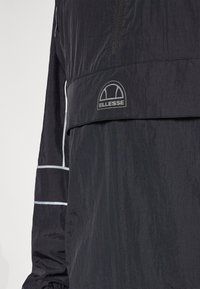 Gros plan sur la manche noire de la veste et la poitrine avec des bandes réfléchissantes blanches et le logo Ellesse sur le panneau avant.