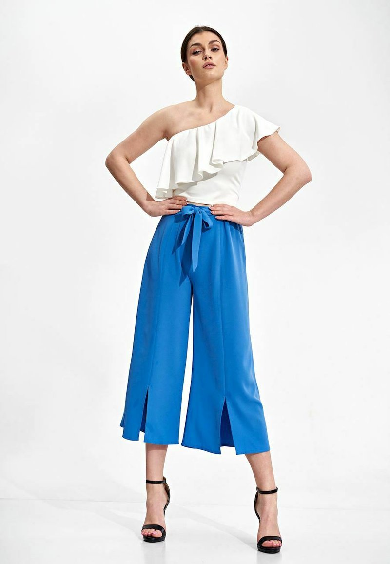 Witte top met één schouder en ruffles, gecombineerd met blauwe cropped broek met wijde pijpen, voorzien van een voorkantopeningen en een strikband in de taille.