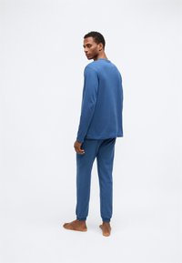 Blå langærmet top og matchende bukser lavet af blødt stof. Bukserne har elastiske manchetter. Modellen står barfodet og er let vendt bagud.