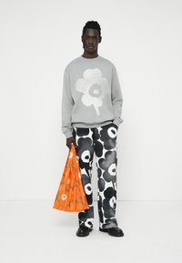 Sudadera gris con gráfico de flores blancas, combinada con pantalones de flores en blanco y negro. El modelo sostiene un bolso floral naranja. Zapatos negros.
