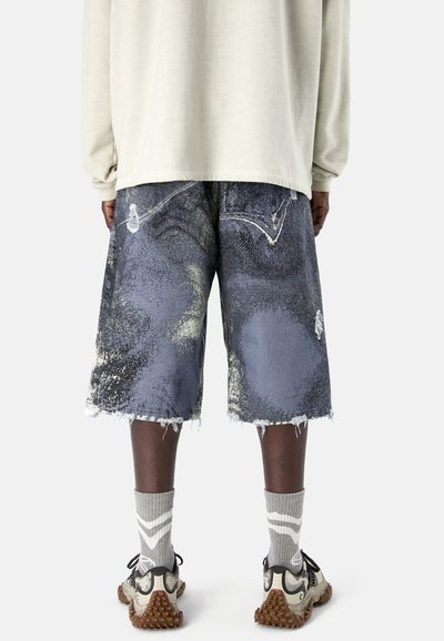 Personne portant un short en denim déchiré arrivant aux genoux avec des ourlets effilochés, des chaussettes gris clair à rayures blanches, et des baskets beiges à semelles texturées.