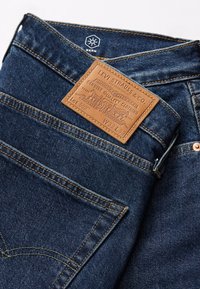 Mörkblå jeans av denim med en brun läderetikett som visar varumärkes- och produktinformation, kopparnitar och kontrasterande sömmar.