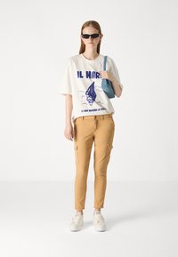 Witte T-shirt met blauwe graphic, beige cargobroek en witte sneakers. Model houdt een lichtblauwe tas vast en draagt zwarte zonnebril.