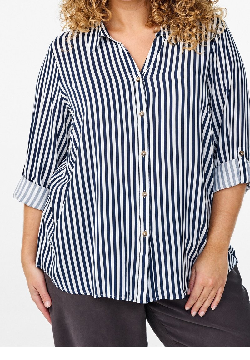 Femme portant une chemise boutonnée à rayures verticales bleu marine et blanche avec manches retroussées et pantalon foncé.