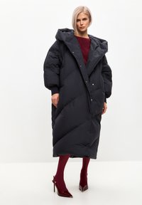 Crna puf jakna s kapuljačom, oversized kroj, dijamantni uzorak, kopčanje na dugmiće, u kombinaciji s burgundskim tajicama i sjajnim crvenim visokim potpeticama.