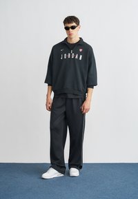 Μαύρο φαρδύ φούτερ με το λογότυπο "JORDAN" σε άσπρα και κόκκινα γράμματα, συνδυασμένο με μαύρο παντελόνι φόρμας με άσπρες ρίγες στο πλάι και άσπρα αθλητικά παπούτσια.