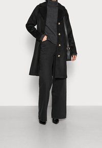 Manteau noir à grands revers et boutons dorés, associé à un col roulé gris foncé et un pantalon noir ample. Petit sac à main noir et chaussures à talons pointues.