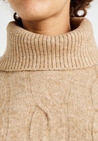 Maglione beige lavorato a maglia con colletto alto a coste e motivo a trecce, che presenta una texture morbida e delle leggere sfumature.