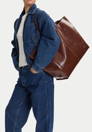 Mujer con chaqueta y pantalones vaqueros azules, camisa blanca, llevando un gran bolso tote de cuero marrón al hombro.