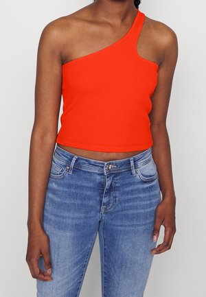 Kvinde iført en lys orange, enkelt skulder crop top kombineret med blå, højttaljede jeans mod en ensfarvet baggrund.