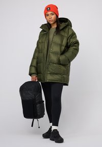 Giubbotto puffer verde oliva con cappuccio, abbinato a leggings neri e sneaker. Zaino nero con finitura testurizzata e dettagli del logo. Berretto arancione.