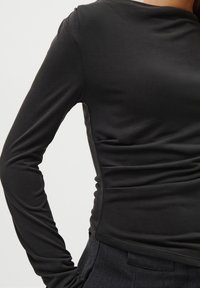 Crna tunika dugih rukava od glatkog, rastezljivog materijala s prilagođenim siluetom i suptilnim naborima sa strane. Nema vidljivih šavova ili metalnih detalja.