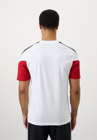 Camisa desportiva de manga curta com um corpo branco, mangas texturizadas em vermelho e riscas laterais pretas. O tecido parece leve e suave.