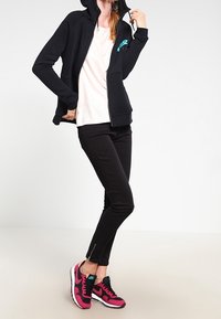 Svart zip-up hoodie med framfickor och turkos logotyp, ihop med en ljusrosa topp, svarta byxor med ankellåsningsdragkedjor och rosa sneakers.