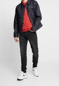 Veste en jean bleu foncé avec des boutons métalliques portée sur un t-shirt rouge vif, associée à un jean noir et des baskets blanches.
