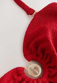 Primo piano di un top bikini in tessuto rosso strutturato con spallina regolabile e chiusura metallica circolare dorata su sfondo chiaro.
