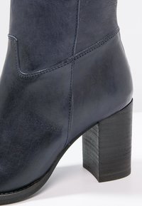 Bottine en cuir bleu marine avec une texture lisse, une forme arrondie et un talon épais en bois avec un grain visible.