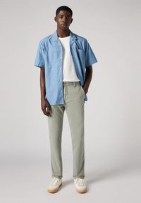 Camicia azzurra a maniche corte, t-shirt bianca sotto, pantaloni verde chiaro e sneakers beige con dettagli bianchi. Tessuto morbido.