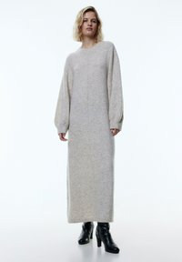Übergroßes, langes graues Strickkleid mit Rundhalsausschnitt und puffigen Ärmeln, strukturierter Stoff, kombiniert mit schwarzen High-Heel-Stiefeln.