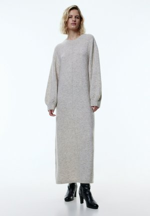 Übergroßes, langes graues Strickkleid mit Rundhalsausschnitt und puffigen Ärmeln, strukturierter Stoff, kombiniert mit schwarzen High-Heel-Stiefeln.