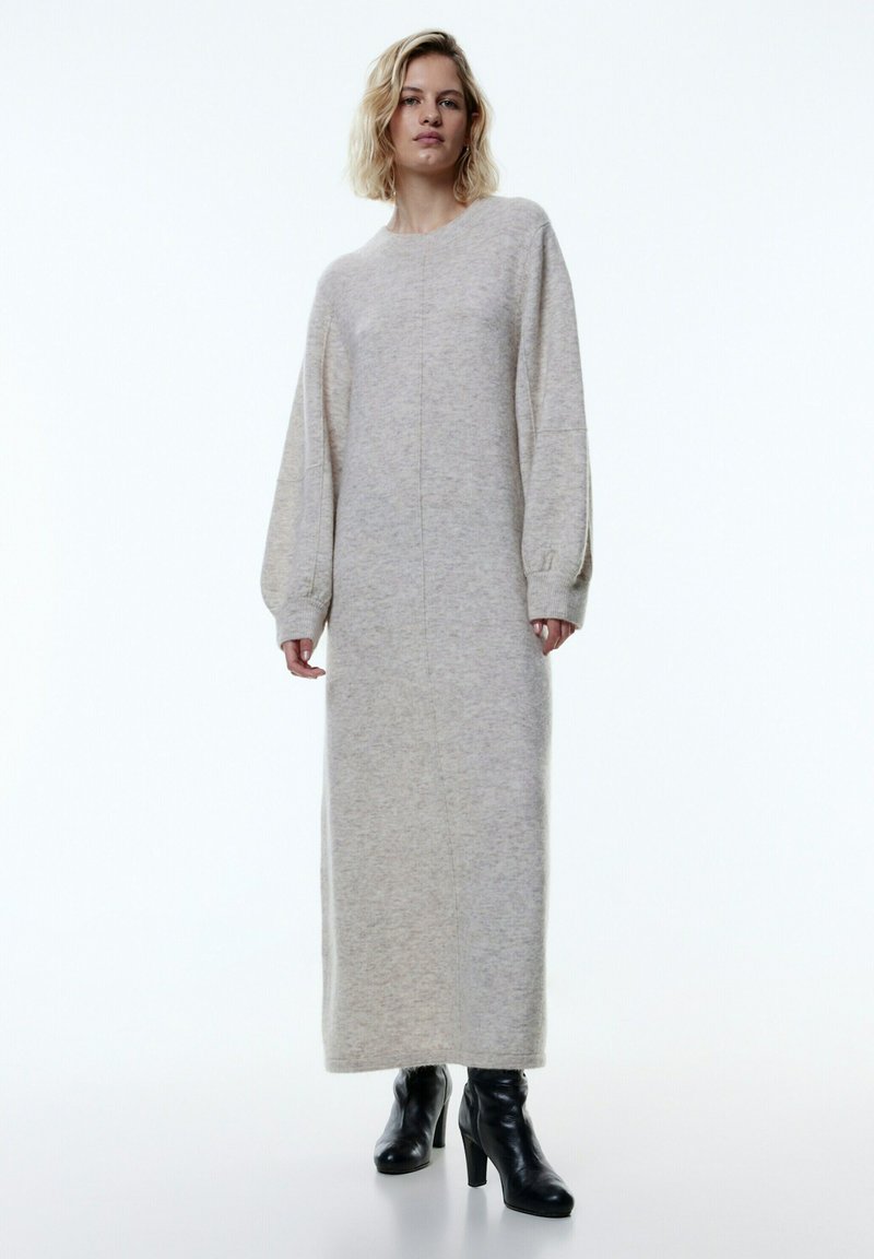 Übergroßes, langes graues Strickkleid mit Rundhalsausschnitt und puffigen Ärmeln, strukturierter Stoff, kombiniert mit schwarzen High-Heel-Stiefeln.