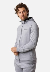 Mies pukeutuneen vaaleanharmaaseen vetoketjulliseen huppariin ja siihen sopiviin housuihin, katsoo sivulle kädet edessä ristissä.